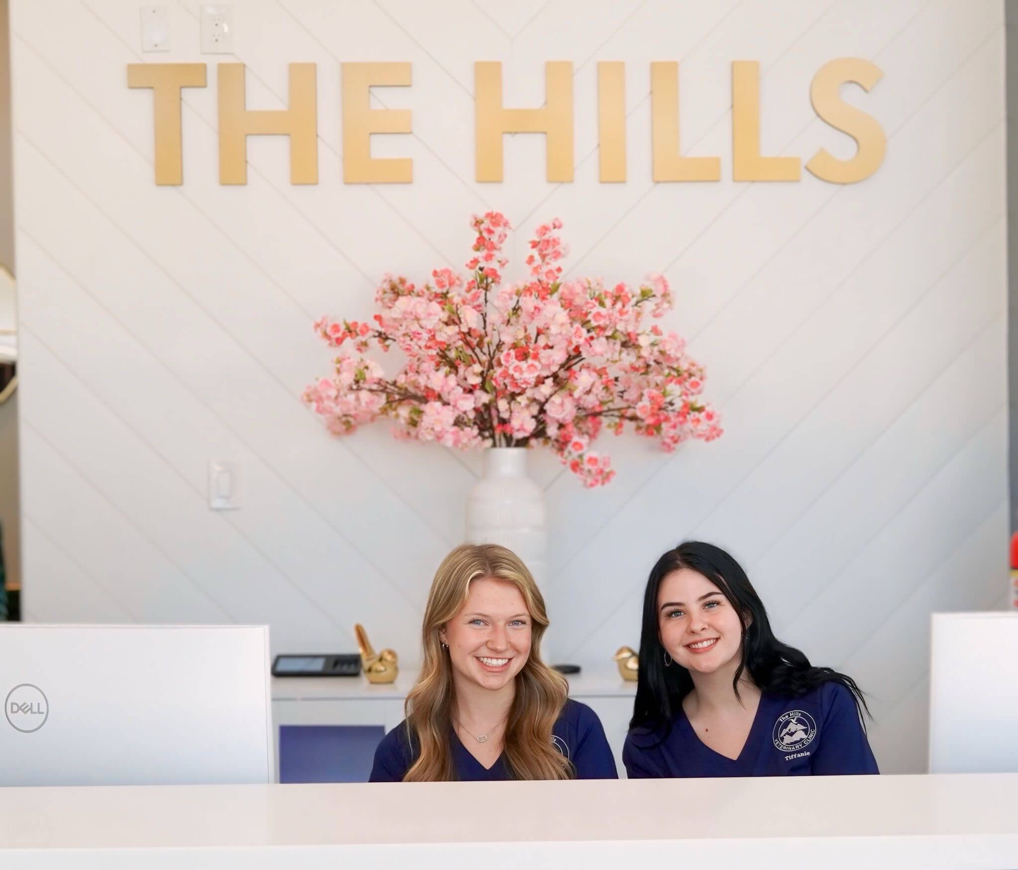 Two women sit behind a white reception desk with pink flowers in a vase and a sign reading "THE HILLS" on the wall behind them.