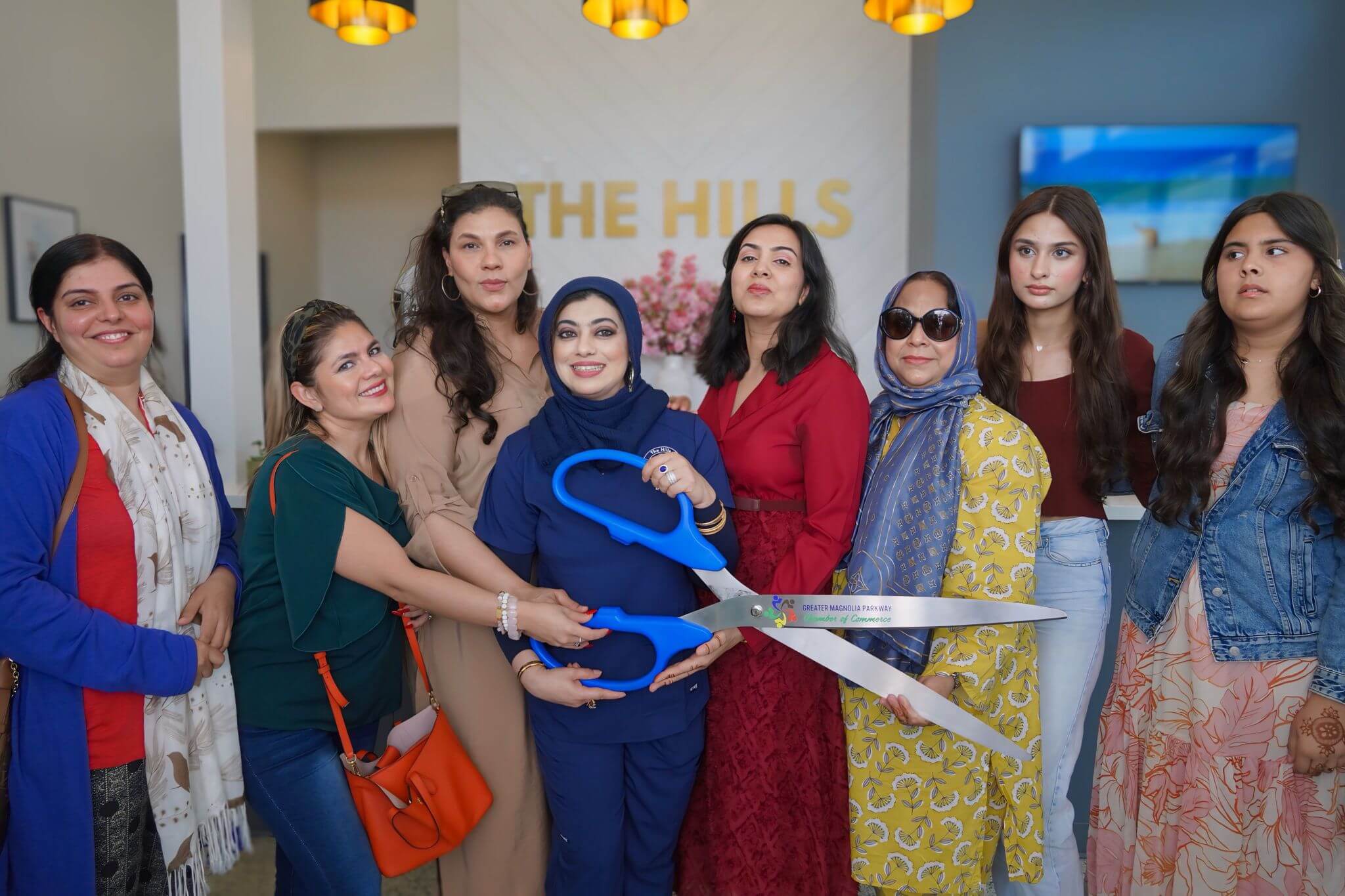 A group of women poses indoors at an event, with one holding oversized ceremonial scissors in front of a sign that reads "THE HILLS.