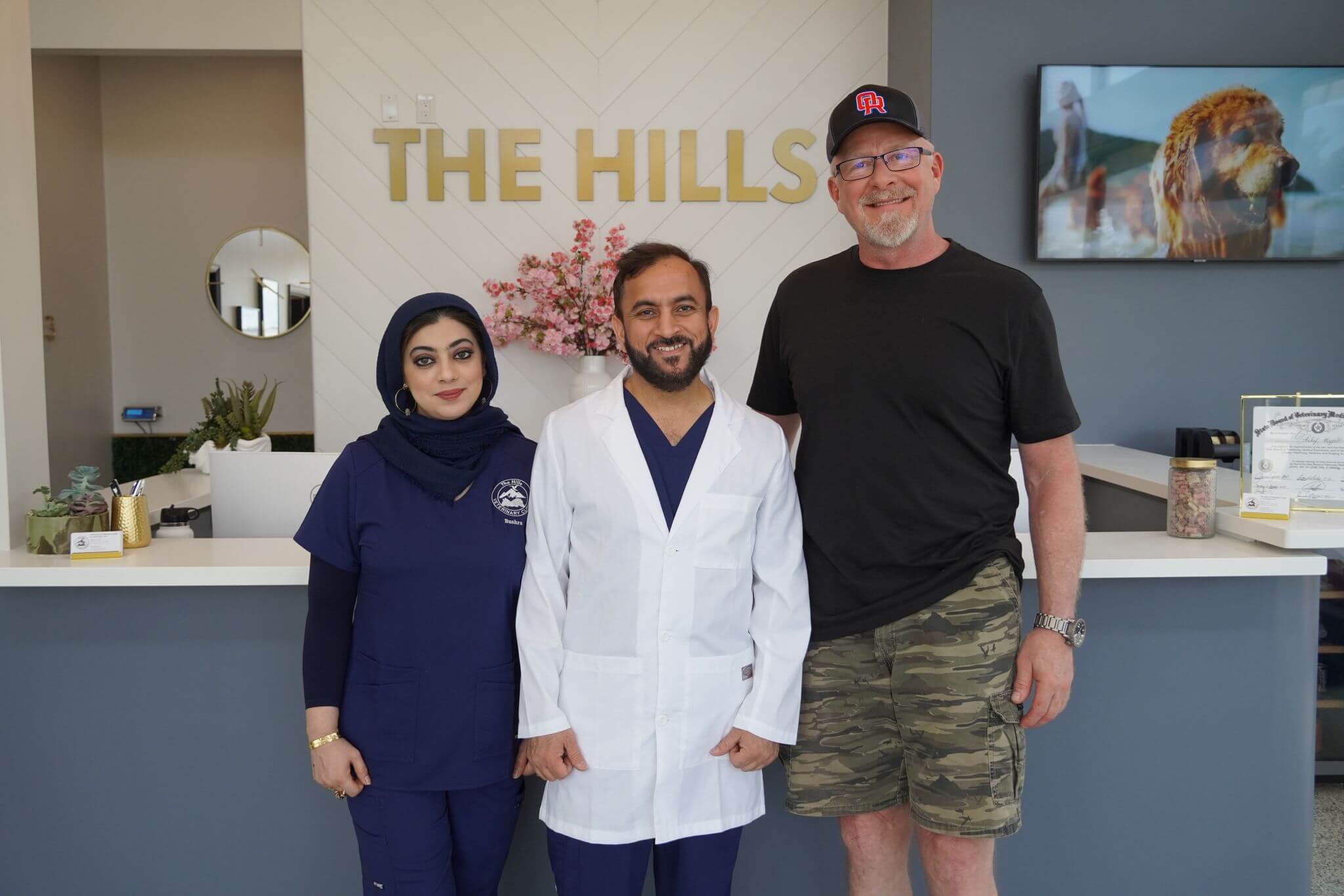 Three people stand together in front of a reception desk labeled "The Hills"; two wear medical uniforms, and one wears casual clothes.