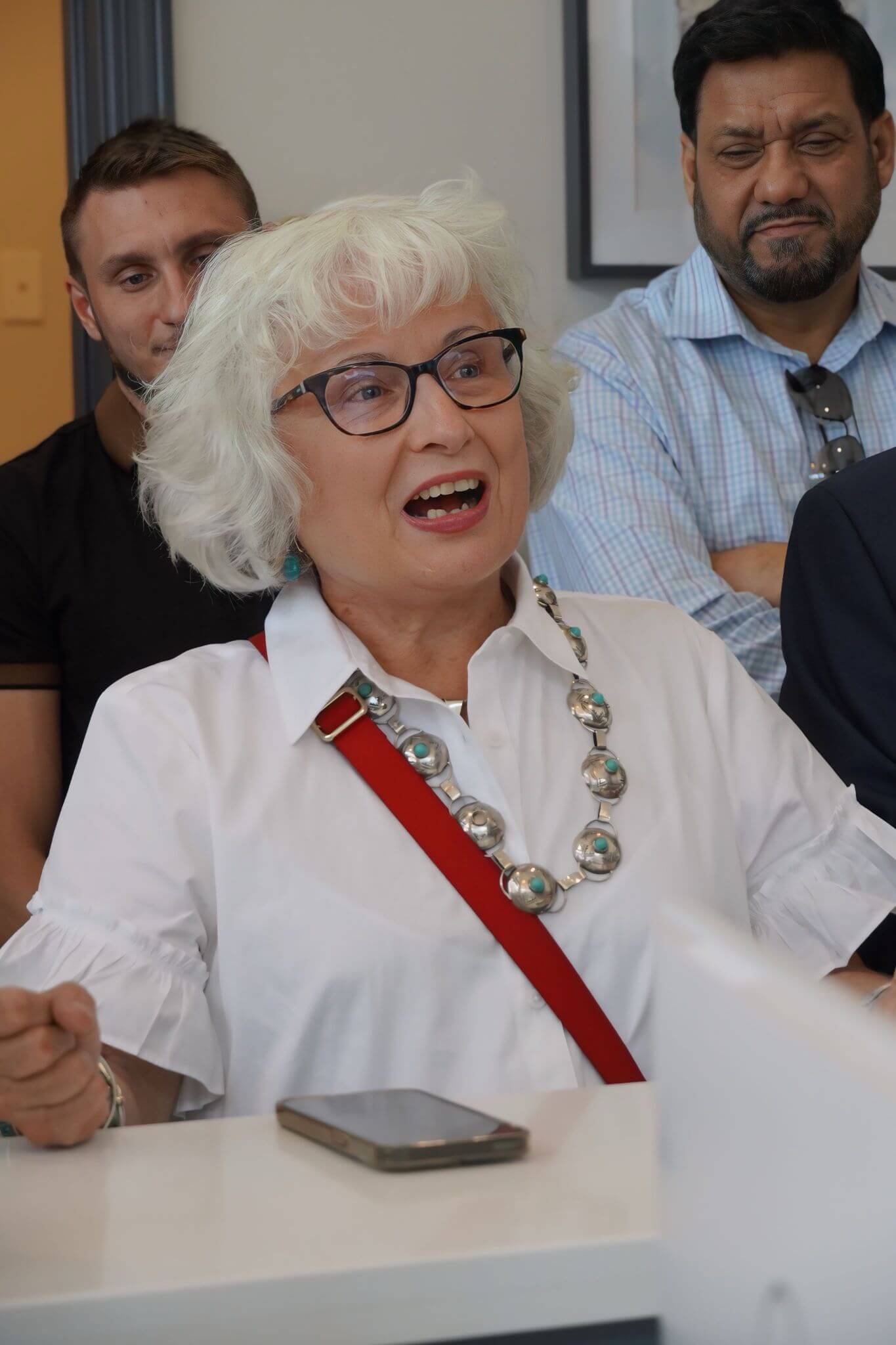 An older woman with white hair and glasses speaks animatedly while seated at a table, with three men standing behind her and a smartphone on the table.