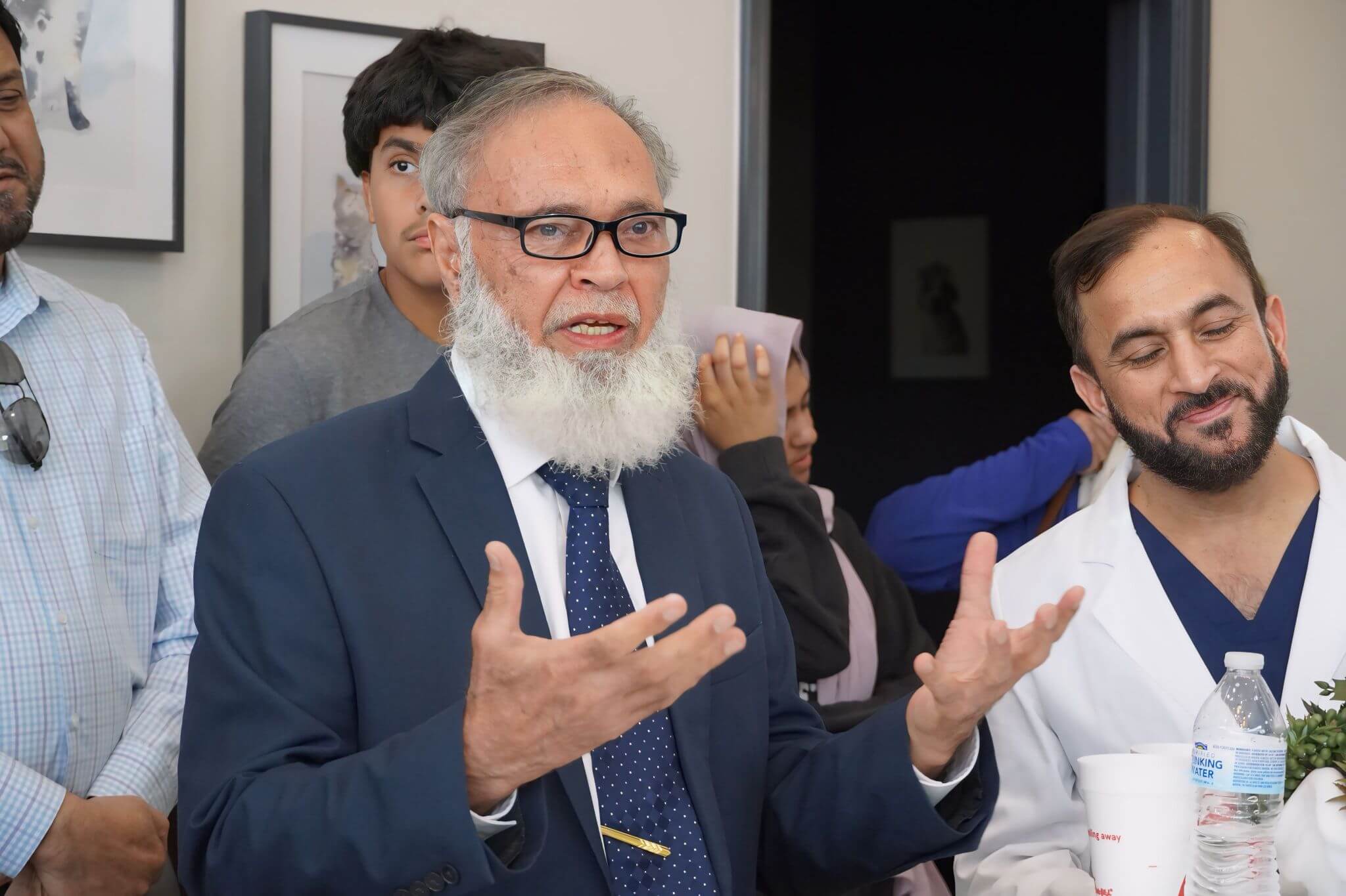 An older man in a suit speaks with hands raised while others, including a man in a white coat, listen in a room.
