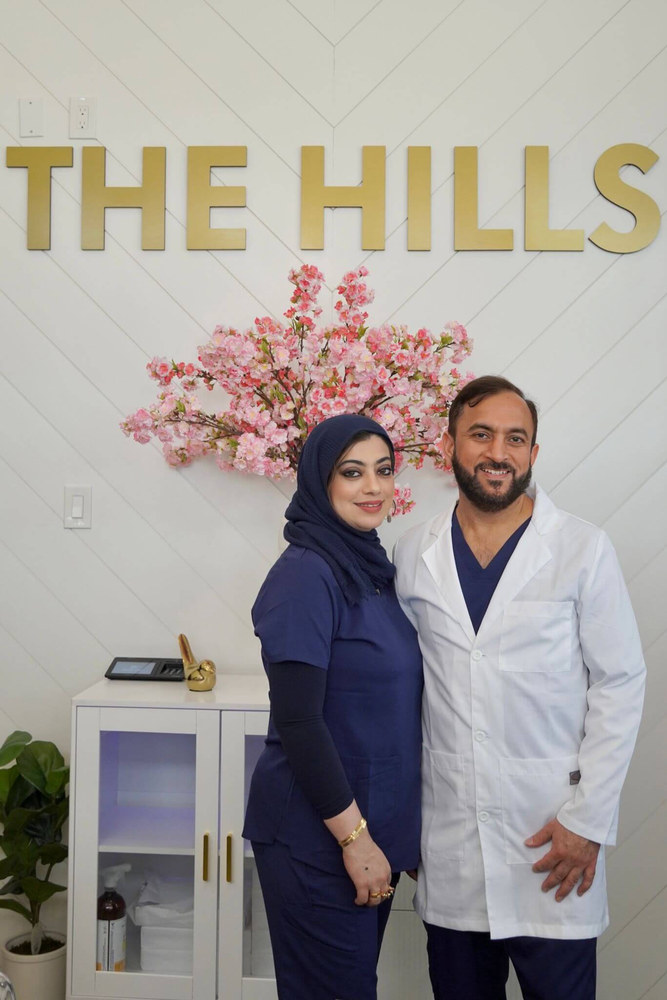 Two medical professionals pose in front of a sign reading "THE HILLS" with pink flowers and a cabinet in the background.